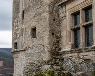 Close-up of an old stone château wall in Lozère showing both sturdy medieval masonry and a later, more refined window detail, under soft overcast light with lichen and moss visible on the stone.
