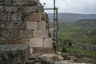 Section of a Lozère castle wall mid-restoration, weathered masonry with scaffolding and chalk marks, plateau landscape in soft morning light, no people, natural subdued colors.