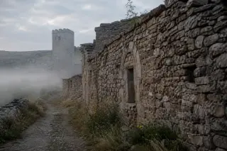 Subtle view along a rural path in Lozère with a layered stone wall, a distant square tower partially hidden by morning mist, and soft light illuminating textured masonry details.